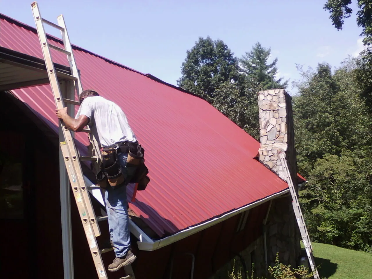 Roofer on a ladder beside a red metal roof and stone chimney — Soffit & Fascia Repair by Roofing Brothers in Holderness